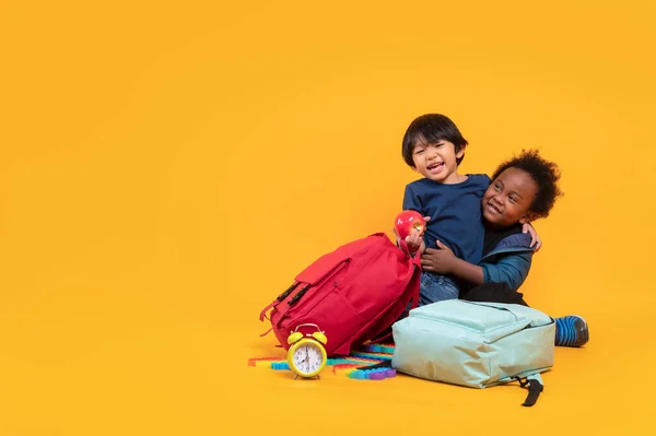 两个3岁的学生 来自不同国籍的亚洲和黑人男孩 带着书包坐在一起 在黄色的背景下独自玩耍 有着复制的空间 国际学生 — 图库照片