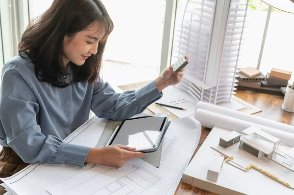 Professional architect women working in designer jobs while using tablet and holding material sample for modern house on the desk having blueprint, Material sample and Building model at loft office