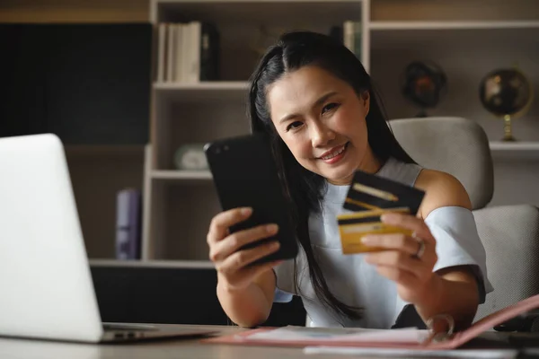 Smile woman holding credit and debit card with smartphone in office front of decoration shelf next to labtop and file