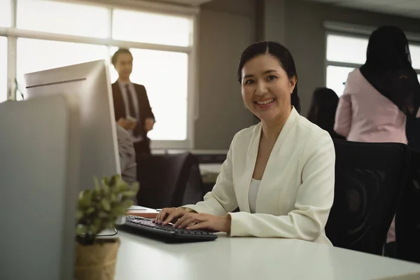 Smiling and confident female employee sit in black chair front of computer was observed by boss next to window in office and many colleague
