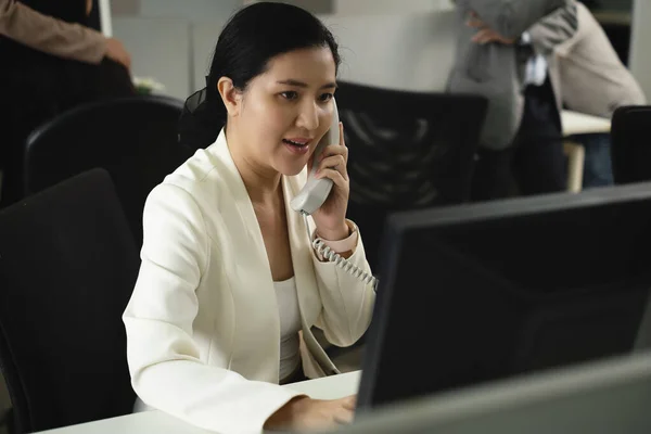 Asian female employee with white suit sit in black chair busy with hotline front of monitor and many queue of co-worker behind in the office