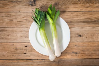 Eski ahşap bir masada beyaz bir tabakta iki taze çiğ pırasa yatıyor. Yukarıdan görüntüle