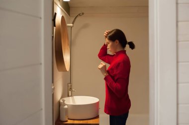 Kırmızı kazaklı genç bir kız evdeki banyoda aynanın önünde saçını tarıyor.