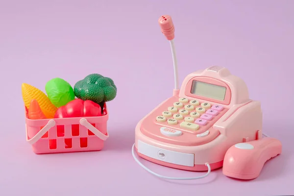 play store, children's toy cash register and basket of vegetables on lilac background, food delivery concept, closeup