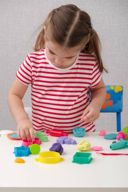 Beyaz ve kırmızı çizgili tişörtlü dikey küçük kız coşkuyla plasticinle oynuyor, hamur oynuyor, oyun hamuru, mankenlik için ayin yapıyor, yaratıcılık için hazırlanıyor.