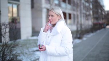 Genç ve çekici bir kadın sokakta sigara içiyor. Beyaz ceketli sarışın, modern sokak modasında.