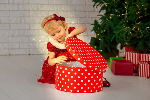 Happy girl opens a Christmas box with a gift. Festive magic for Christmas or New Year. Child near a decorated Christmas tree with lights