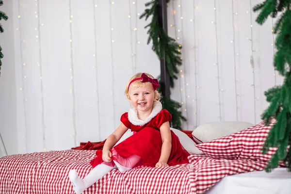 happy playful girl sits on bed decorated with branches of Christmas tree. child woke up on Christmas morning and is waiting for gifts