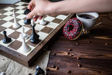 El beyaz bir bardak kahve tutuyor. Satranç tahtası ve dağınık satranç tahtası ahşap masada. Sabah kahvaltısı için çikolatalı çörek. oyun kavramı