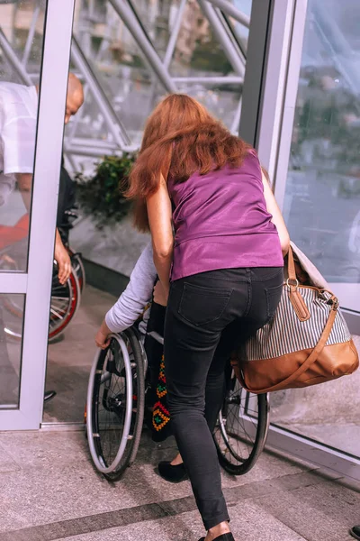 A woman helps a girl in a wheelchair to enter the doors of a shopping center. An assistant to a person with disabilities carries a wheelchair