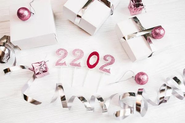 Happy New Year 2022 decorate. Gifts in white boxes with silver wrapping ribbons and pacific pink Christmas balls on a light wooden table.
