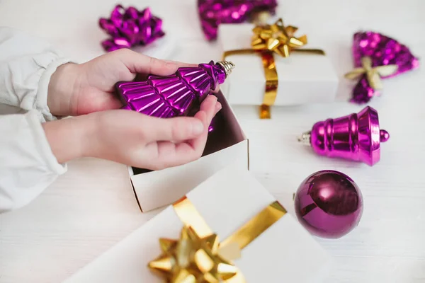 girl holds velvet violet color 2022 year Christmas tree toy in hands . New Year Trend Color 2022 over gift boxes with gold ribbons