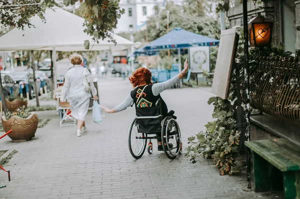 girl in wheelchair rides with hands to side through old town on sidewalk near cars. Freedom of movement of persons with special needs.