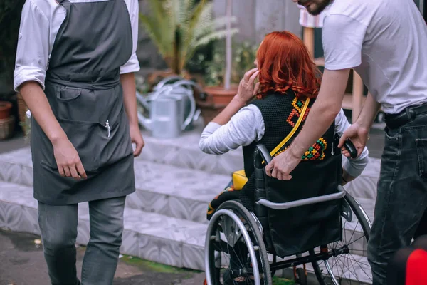 Waiters lift a girl in a wheelchair up the steps without a ramp. A woman is going to have lunch in a cafe inaccessible to people with special needs.