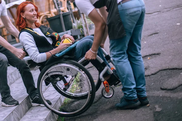 Waiters lift a girl in a wheelchair up the steps without a ramp. A woman is going to have lunch in a cafe inaccessible to people with special needs.