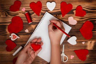 Hands writing down Valentine's Day greetings in a blank notebook. Felt red hearts on a wooden table