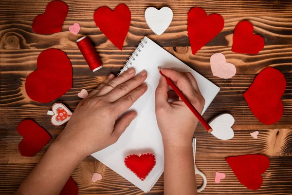 Hands writing down Valentine's Day greetings in a blank notebook. Felt red hearts on a wooden table