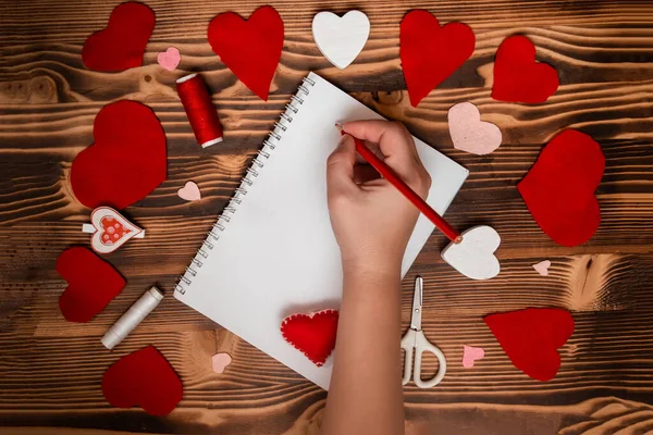 Hands writing down Valentine's Day greetings in a blank notebook. Felt red hearts on a wooden table