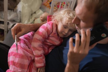 Babası yüksek sıcaklığı termometreyle ölçüyor. Çocuğun ateşi yüksekti. Babamda termometre var. Yatak odasında pijamalı hasta kıza sarılıyor.