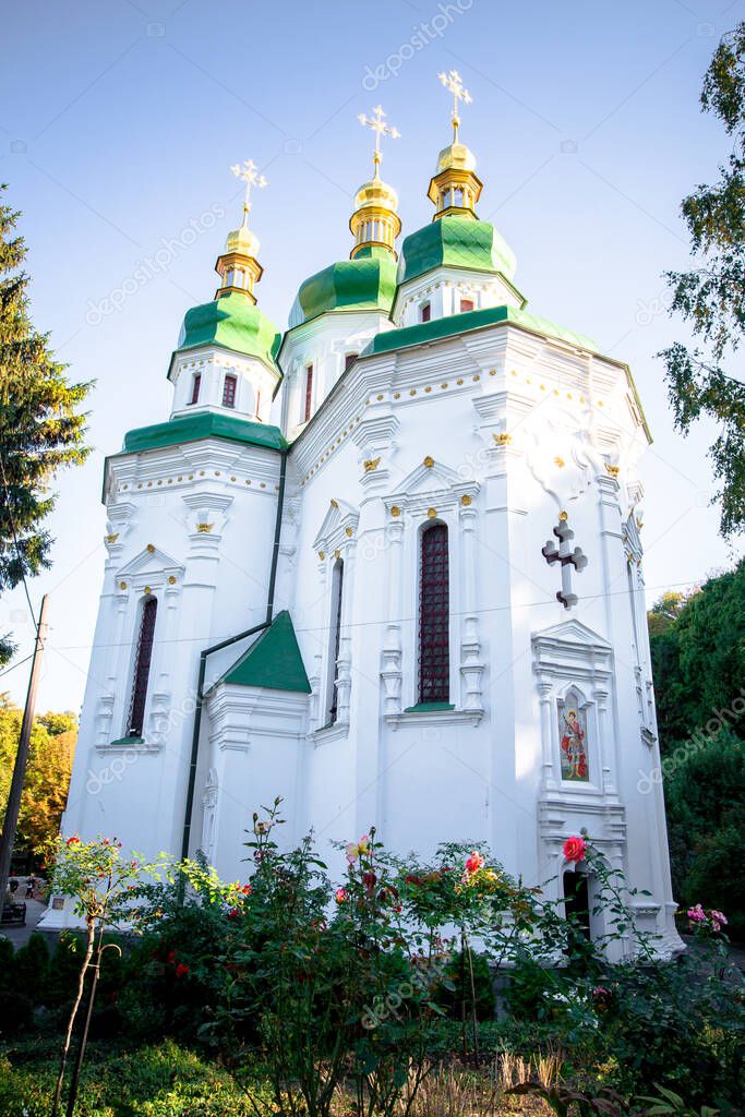 Church of St. George in Vydubitsky Monastery in the city of Kiev. Flowerbeds and trees in the courtyard of the monastery of the forest in the botanic garden. The sunset sun breaks through the leaves.
