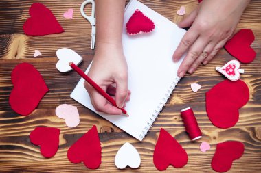Hands writing down Valentine's Day greetings in a blank notebook. Felt red hearts on a wooden table