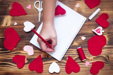 Hands writing down Valentine's Day greetings in a blank notebook. Felt red hearts on a wooden table