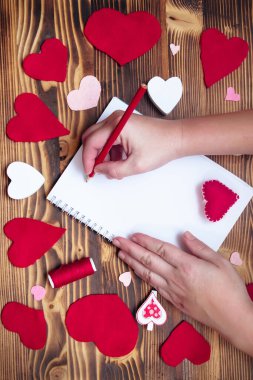 Hands writing down Valentine's Day greetings in a blank notebook. Felt red hearts on a wooden table