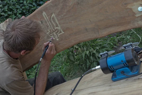 man with device cuts out coat of arms of Ukraine. man cuts picture on tree with Drill for wood carving. man cuts an ornament with cutter - nozzle on drill