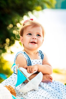 Bir çocuk ağaçların yanında sahilde oturuyor. Küçük kız ayaklarını çapraz yaptı ve elini ağzının yanında tuttu. Oyuncak tekne tutan çocuk. Komik kız ebeveynlerine tökezler..