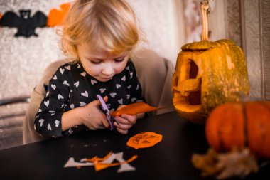 Cadılar Bayramı için çocuk aktivitesi. Bir çocuk ev için turuncu kağıt süslemeleri kesiyor. Tatil akşamından önce ev hayatı.