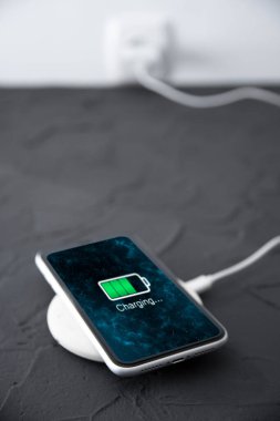 Koyu arkaplandaki kablosuz şarj cihazında cep telefonu var. Ekranda simge bataryası ve şarj süreci aydınlatması. Güç kaynağına bağlı akıllı telefonlar. Düşük pil seviyesi sorunları..