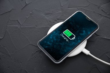 Koyu arkaplandaki kablosuz şarj cihazında cep telefonu var. Ekranda simge bataryası ve şarj süreci aydınlatması. Güç kaynağına bağlı akıllı telefonlar. Düşük pil seviyesi sorunları..