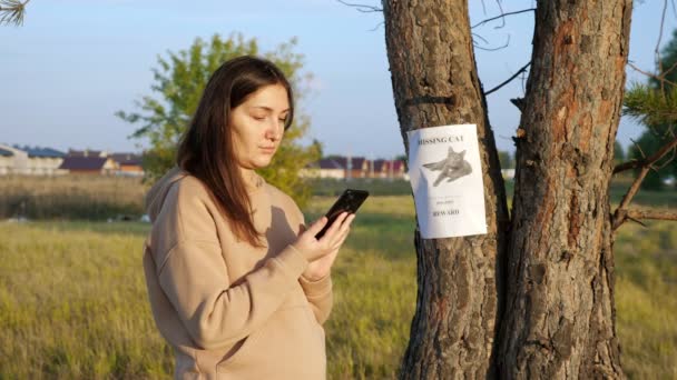 Vrouw in hoodie wijzerplaten nummer van vermiste kat poster in park — Stockvideo