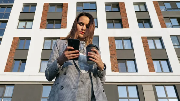 Egy öltönyös nő kávét iszik egy műanyag pohárból, és belenéz a telefonba egy épület hátterében. — Stock Fotó