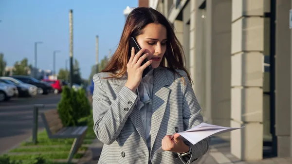 Fiatal nő egy üzleti öltönyben beszél telefonon, és nézi dokumentumok séta az utcán — Stock Fotó