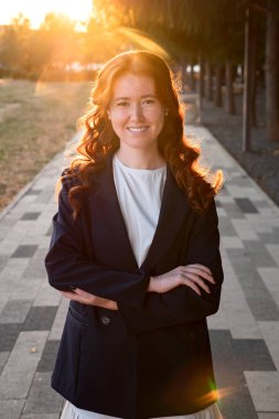 Takım elbiseli, kıvırcık kızıl saçlı, parkta gülümseyen genç bir kadın portresi.