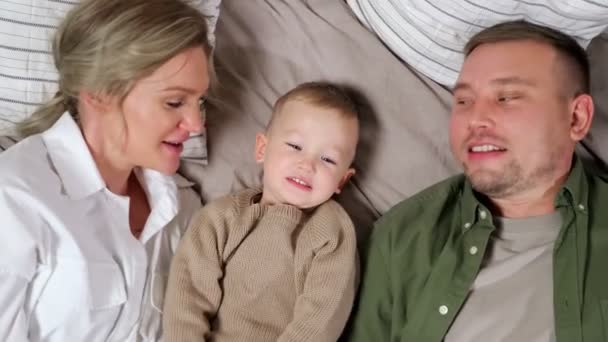 Отец и мать целуют маленького сына, лежащего посреди кровати — стоковое видео
