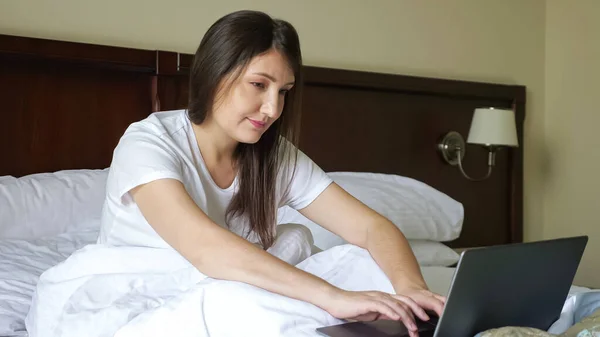 Jovem mulher digitando em um laptop enquanto sentado na cama coberto com um cobertor — Fotografia de Stock