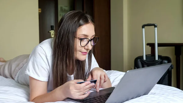 Mulher em óculos com um cartão funciona em um laptop, deitado na cama no estômago contra o fundo de uma mala — Fotografia de Stock