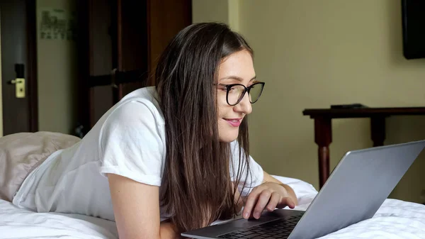안경을 쓴 채웃고 있는 젊은 여자가 침대 위에 누워 있는 노트북을 보고 있습니다. — 스톡 사진
