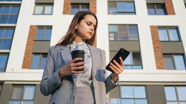 Egy fiatal nő üzleti ruhában ránéz a telefonra és kávét iszik egy műanyag pohárból egy épület hátterében. — Stock Fotó