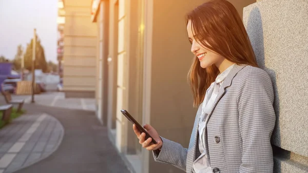 Mladá žena v obleku se podívá na telefon na ulici v blízkosti budovy — Stock fotografie