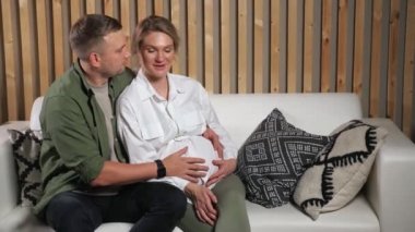 Adam beyaz koltukta oturan hamile kadının karnını okşar.