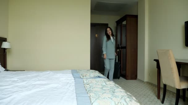 Vermoeide vrouw in een zakelijk grijs pak met een koffer komt een hotelkamer binnen en valt op het bed — Stockvideo
