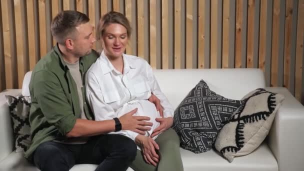 Mann streichelt Schwangeren auf weißem Sofa sitzend den Bauch — Stockvideo