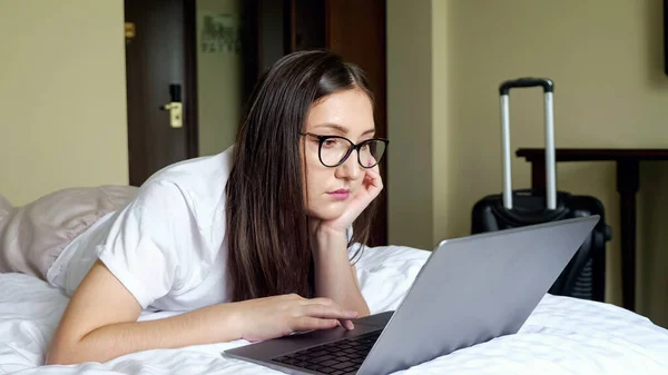 Jovem mulher em óculos olha para um laptop deitado na cama no estômago contra o fundo de uma mala — Fotografia de Stock