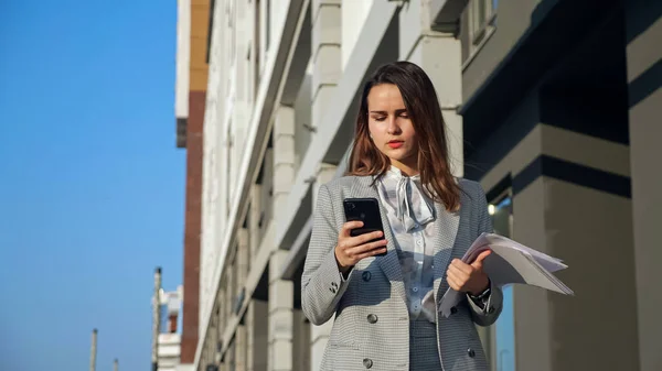 Fiatal nő egy üzleti öltönyben egy telefonnal és dokumentumok sétálnak az utcán — Stock Fotó