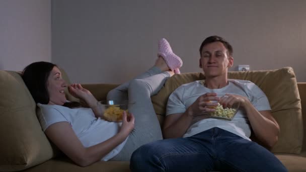 Мужчина бросает попкорн даме, лежащей на диване, смотря телевизор — стоковое видео