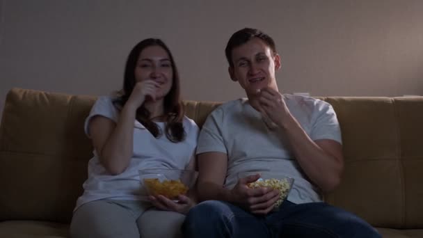 Мужчина с попкорном и женщина с чипсами смеются перед телевизором — стоковое видео