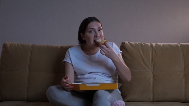 Pani bierze kawałek pizzy i je oglądając telewizję na kanapie — Wideo stockowe